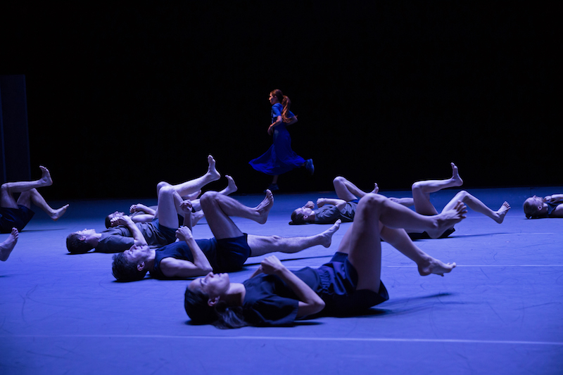 A woman in a blue dress runs in the background as a group of dancers in black costumes lay on the floor with their legs in the air